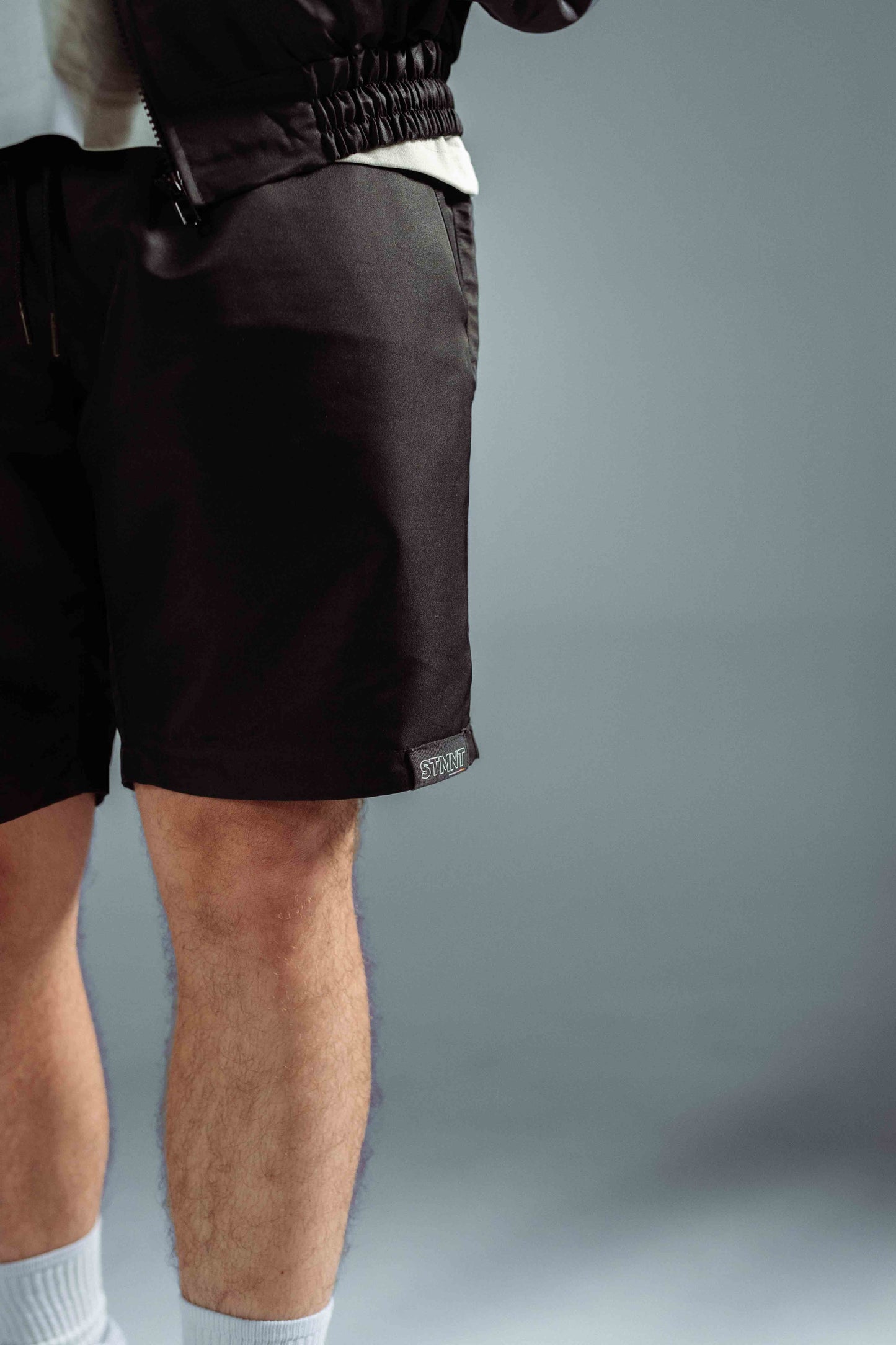STMNT Wear First Edition black athletic shorts side view showing elastic waist, hem STMNT logo patch, and jacket pairing detail, studio shot