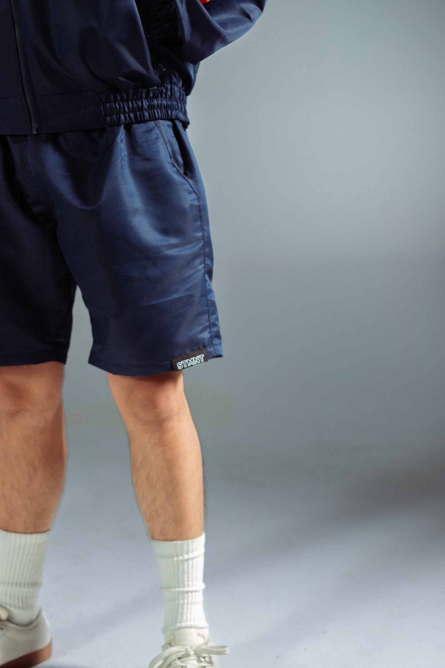 STMNT Wear deep navy athletic shorts side view showing elastic waist, hem STMNT logo patch, and matching jacket pairing, studio shot