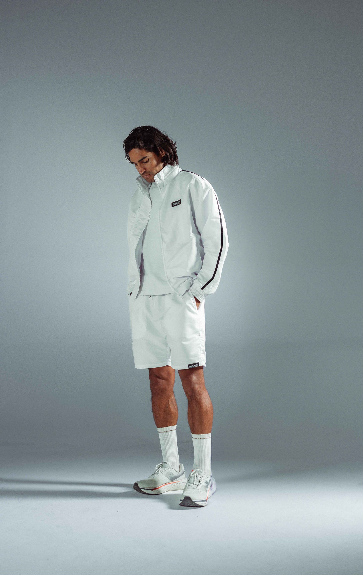 STMNT Wear White Full Tracksuit - zip-up track jacket with dark stripe and matching shorts, model looking down in studio