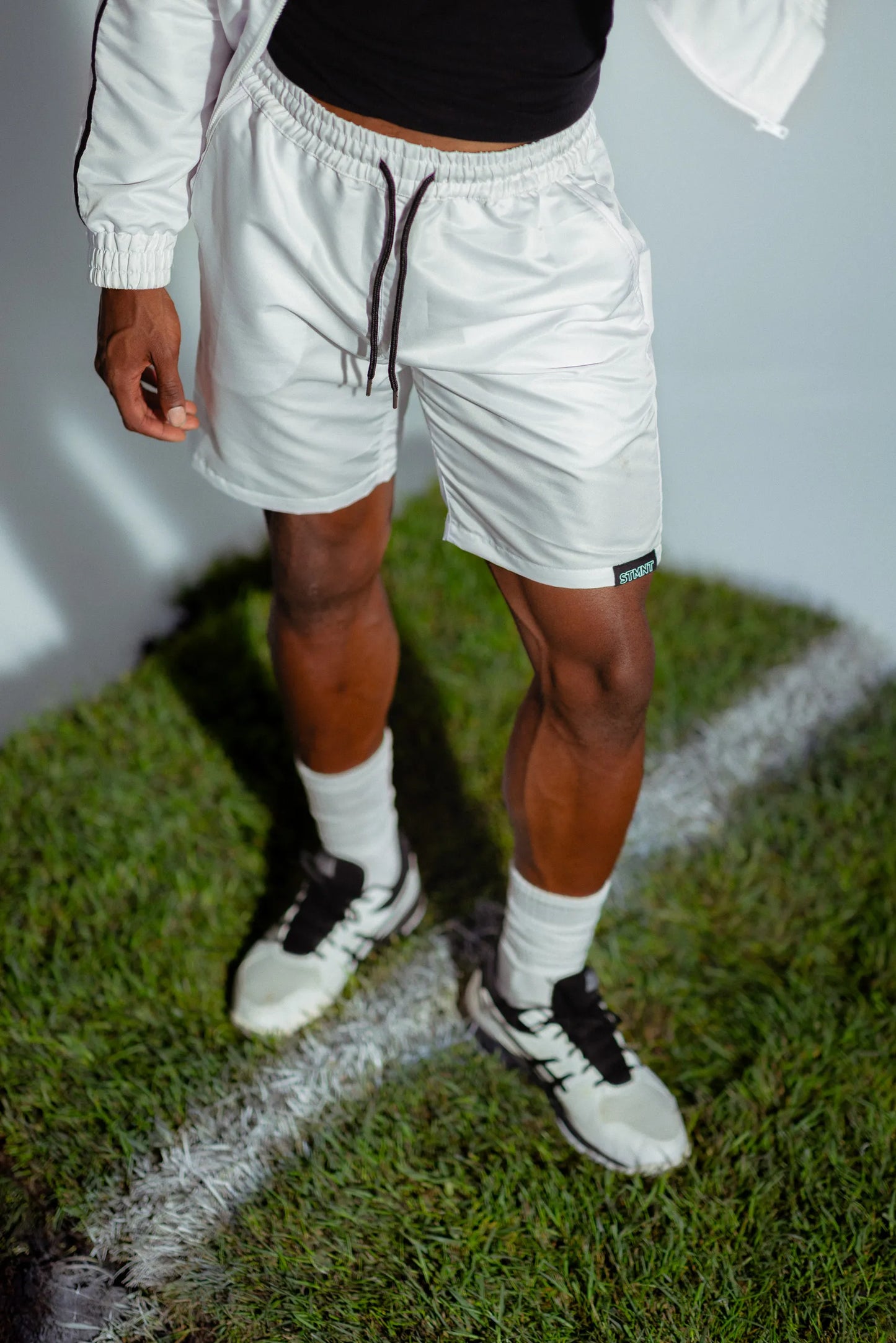 STMNT Wear white drawstring shorts close-up with logo patch, model in football boots on pitch grass