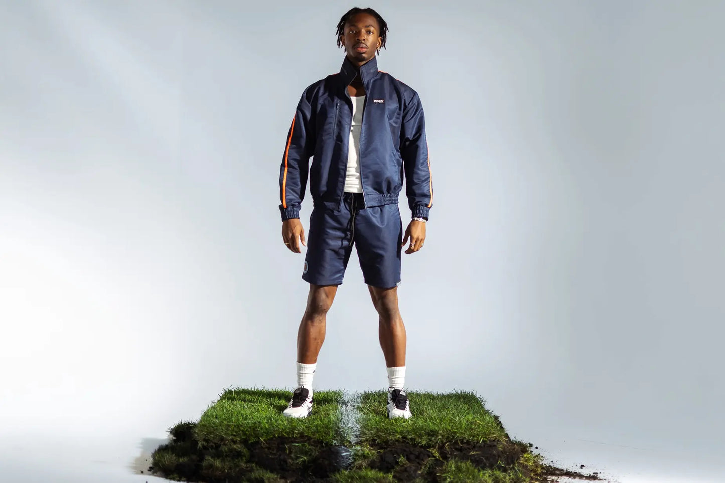 STMNT Wear deep navy full tracksuit on model standing on soccer pitch turf cut-out inside the studio, orange stripe detail