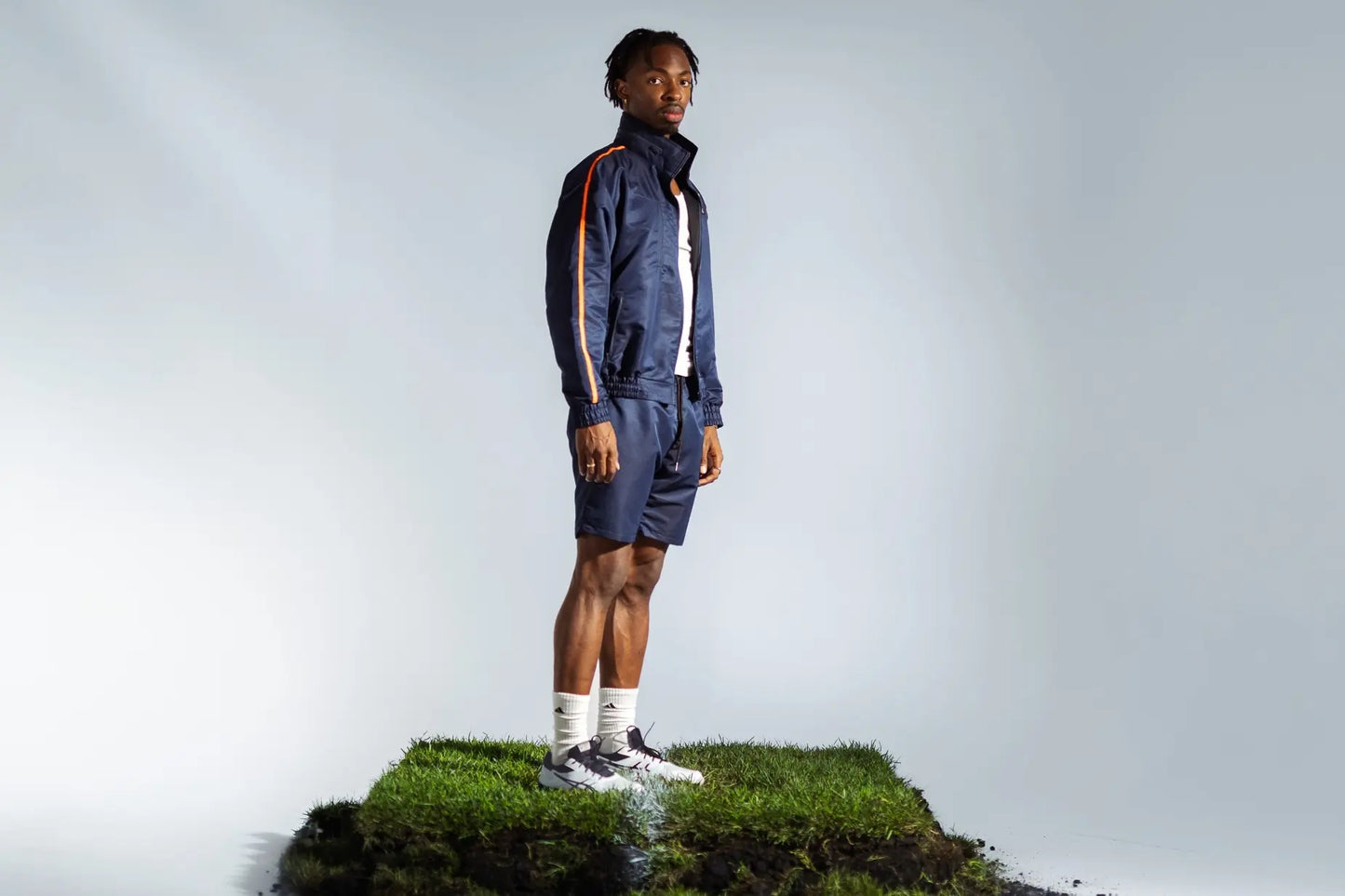 STMNT Wear Deep Navy Full Tracksuit - zip-up track jacket and shorts with orange accent stripe, model on grass pitch turf cut-out in a studio