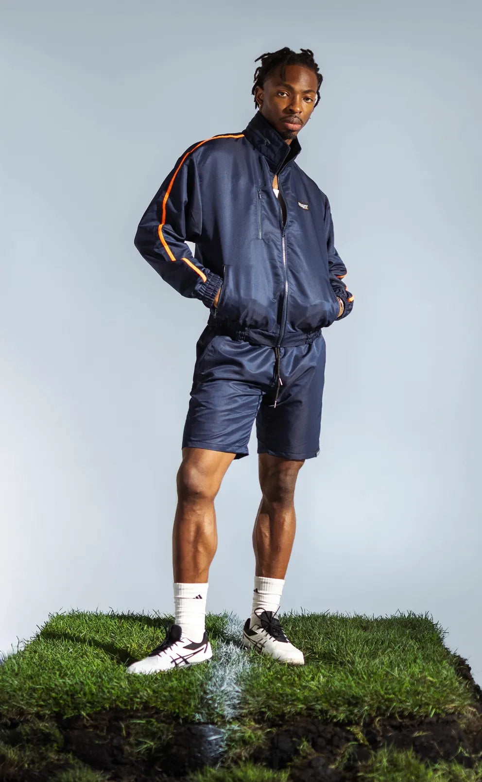 Model wearing STMNT Wear deep navy shorts and matching track jacket on soccer pitch cut-out with football cleats, a studio shot
