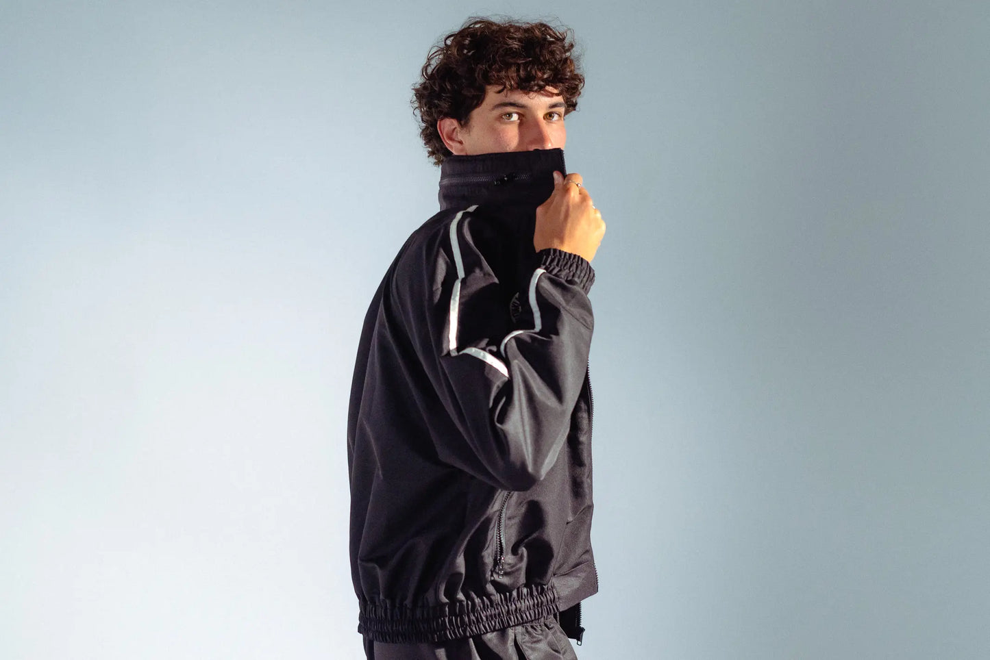 Model zipping up STMNT Wear black track jacket with high collar and white stripe detail, side profile