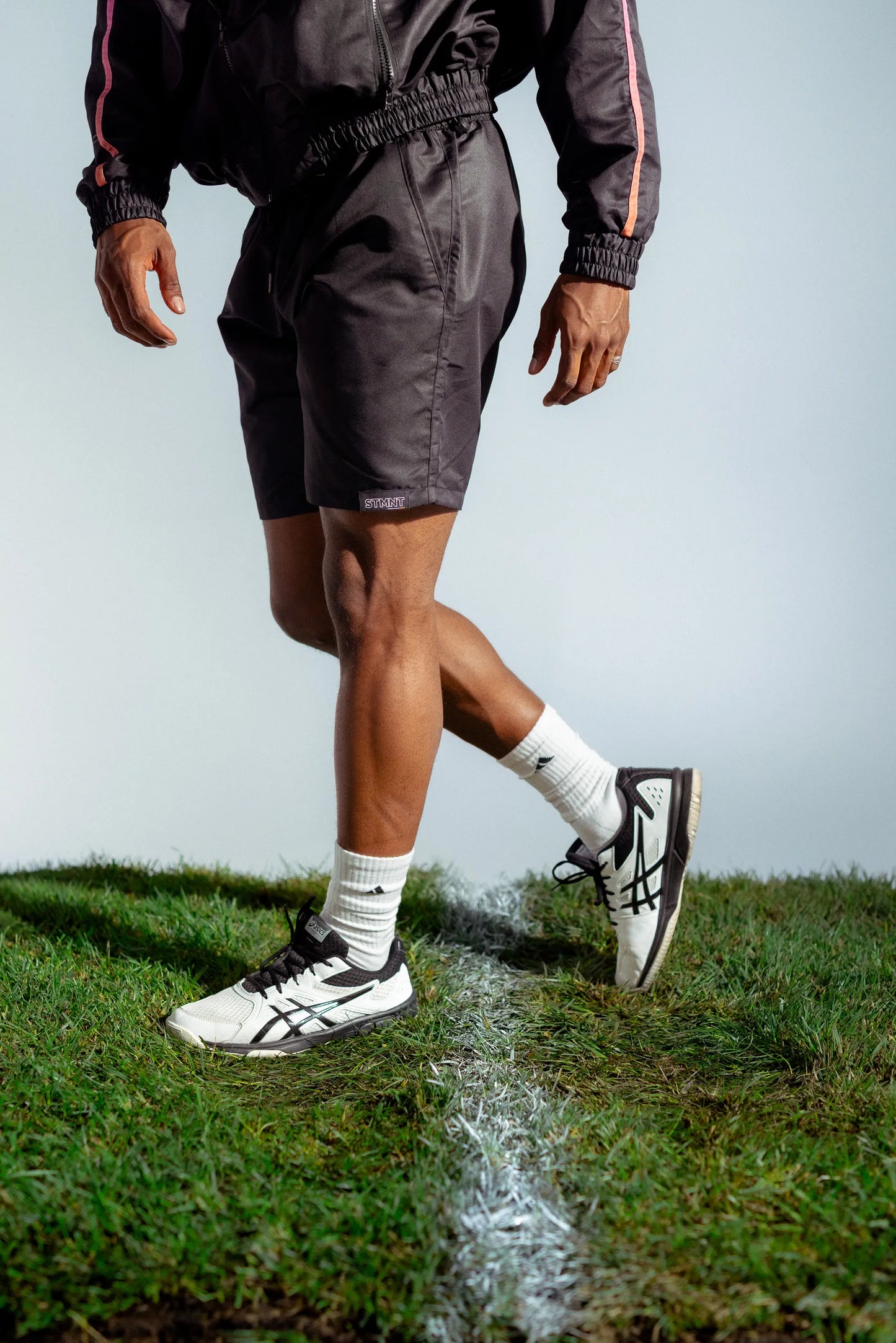 Lower body shot of STMNT First Edition Wear black athletic shorts with jacket walking on soccer pitch grass cut-out in a studio