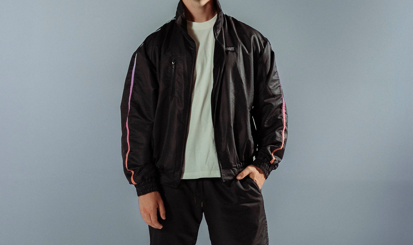 STMNT Wear First Edition black track jacket with gradient orang-blue sleeve stripes open over white tee, studio front view