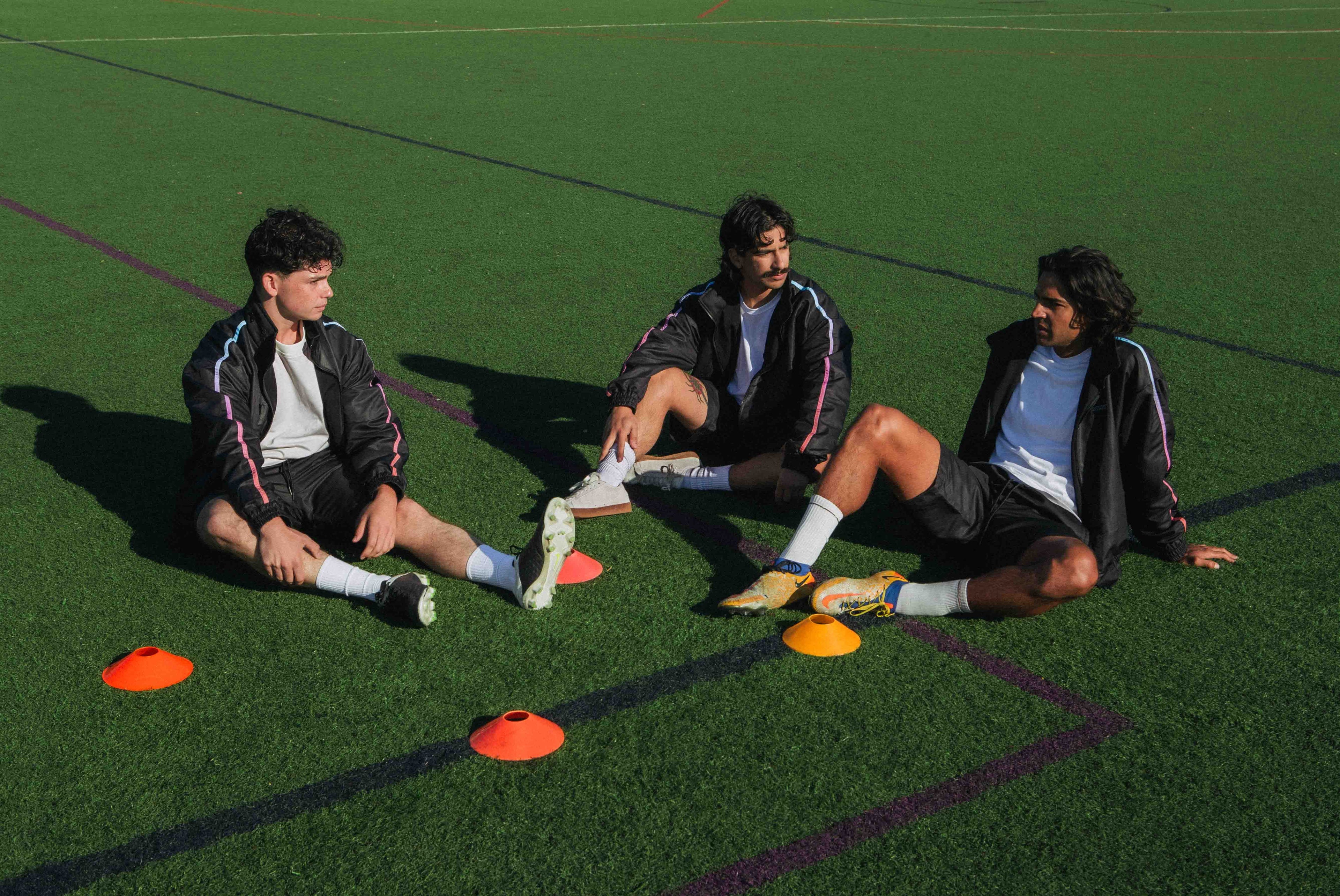 Three models in STMNT Wear black track jackets and shorts relaxing on soccer pitch with training cones and cleats