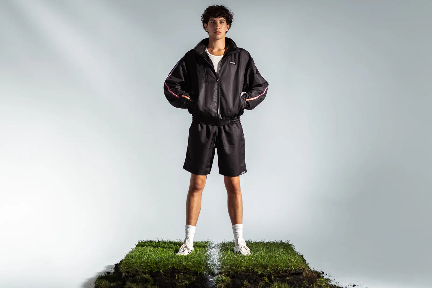 STMNT Wear First Edition black track jacket with orange-blue gradient accent stripes, full outfit on model standing on soccer turf cut-out in a studio