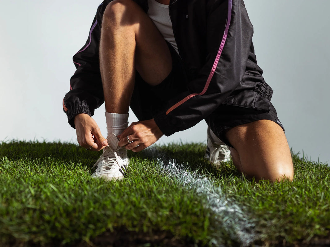 Model tying football boots on grass pitch wearing STMNT Wear First Edition black jacket with orange-pink sleeve trim