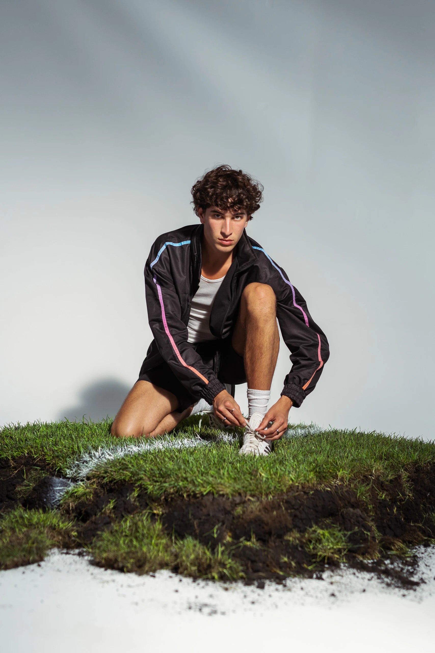 Model crouching on a piece of grass pitch tying cleats in STMNT Wear Firs Edition black track jacket and shorts - soccer-ready athleisure