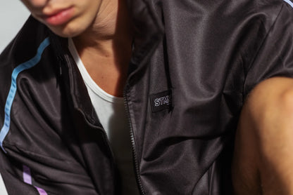 Close-up of STMNT Wear woven chest label on First Edition black track jacket with blue and purple sleeve stripes