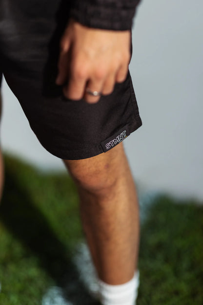 STMNT Wear First Edition black athletic shorts close-up showing woven hem logo STMNT label, model on soccer pitch turf with white sneakers