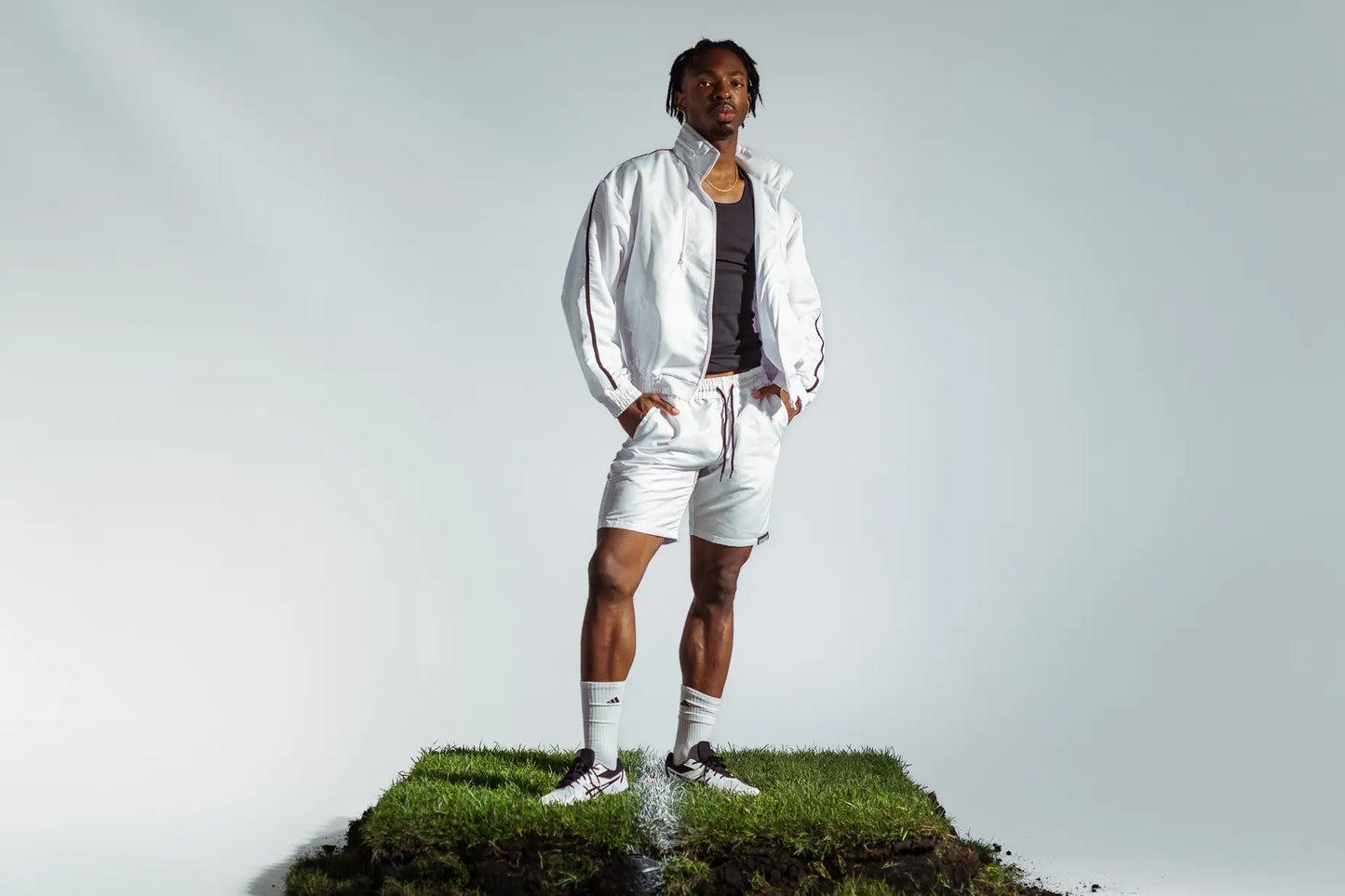 STMNT Wear White Full Tracksuit - track jacket and drawstring shorts over black tank, model in football boots on grass pitch turf cut-out in a studio