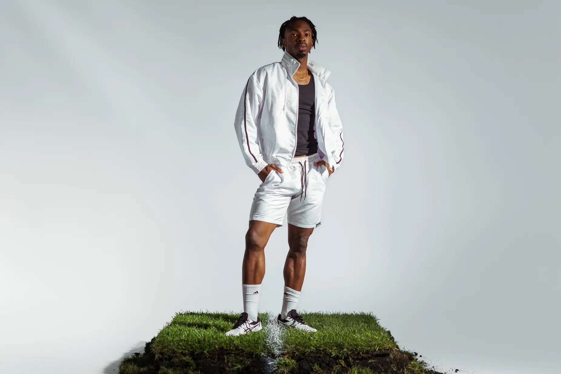 STMNT Wear White Full Tracksuit - track jacket and drawstring shorts over black tank, model in football boots on grass pitch turf cut-out in a studio