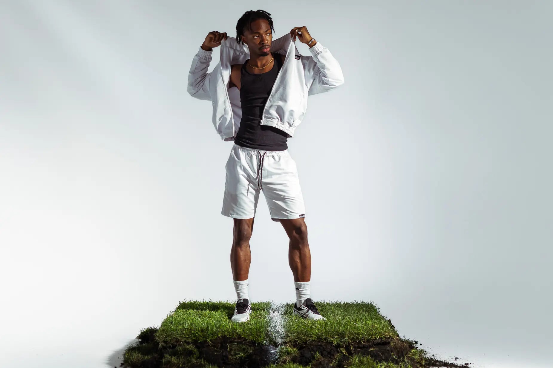 STMNT Wear white track jacket and shorts full tracksuit, model pulling up collar on soccer pitch turf with black tank