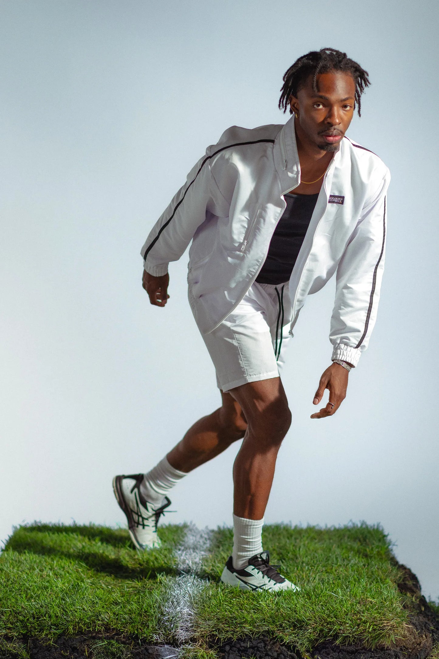 Model in motion wearing STMNT Wear white track jacket and shorts full tracksuit on grass, athletic running pose