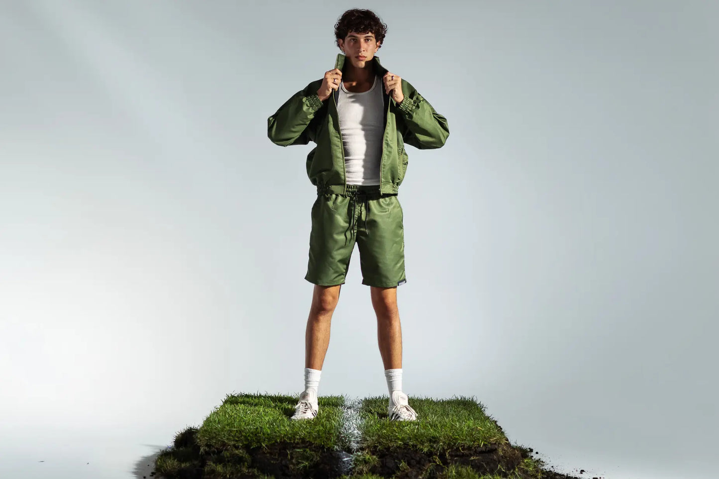 STMNT Wear olive green track jacket and shorts full tracksuit outfit, model pulling collar on soccer turf set