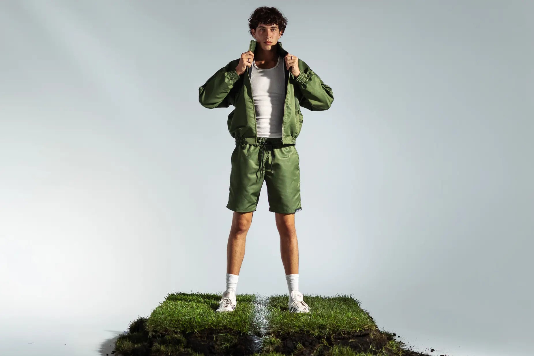 STMNT Wear olive green track jacket and shorts full tracksuit outfit, model pulling collar on soccer turf set