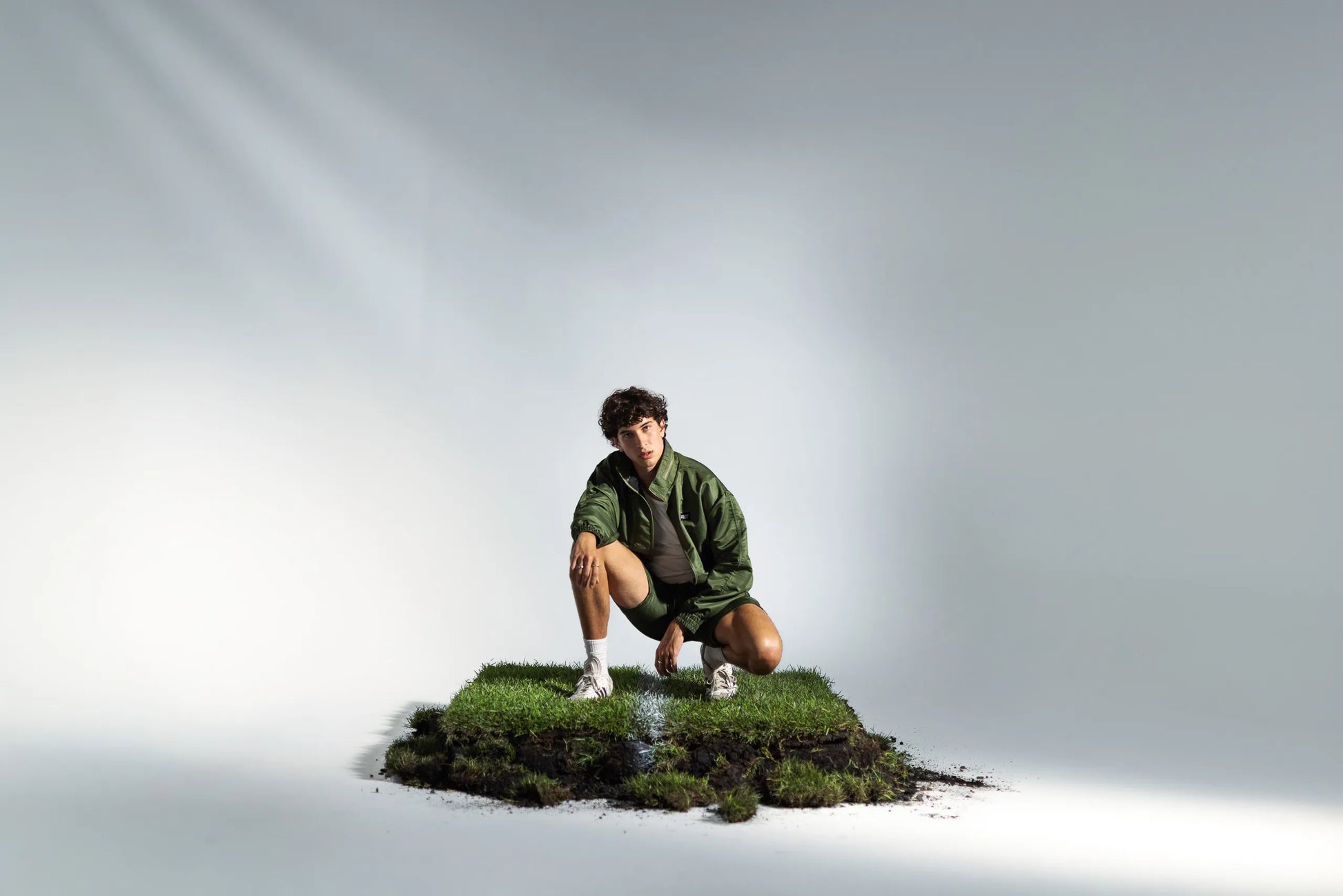 Model crouching in STMNT Wear green athletic shorts and jacket full tracksuit on soccer pitch turf cut-out in a studio