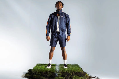 STMNT Wear deep navy full tracksuit on model standing on soccer pitch turf cut-out inside the studio, orange stripe detail
