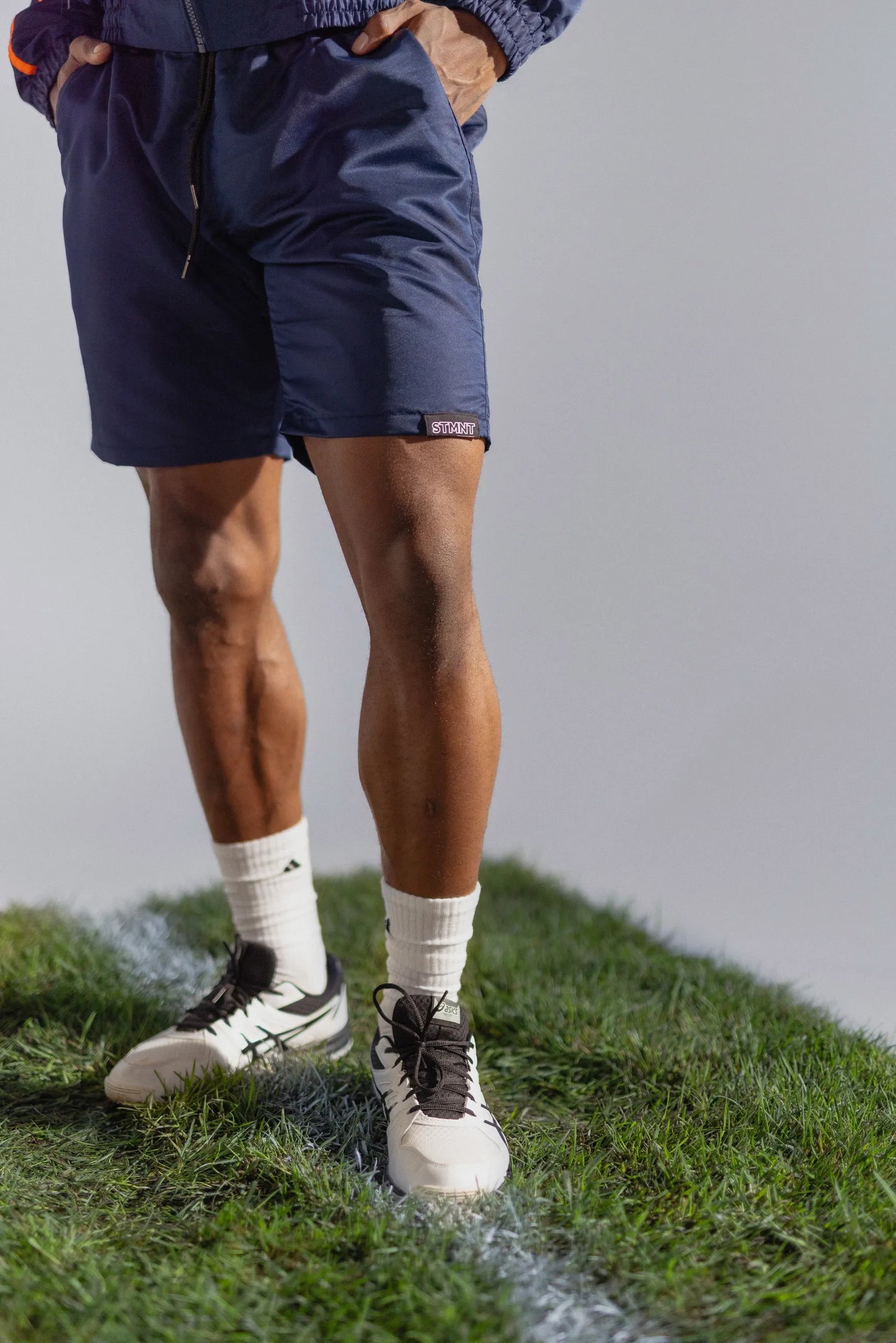 STMNT Wear deep navy athletic shorts close-up with logo label, model in football boots on pitch grass cut-out in a studio
