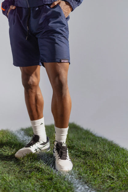 STMNT Wear deep navy athletic shorts close-up with logo label, model in football boots on pitch grass cut-out in a studio