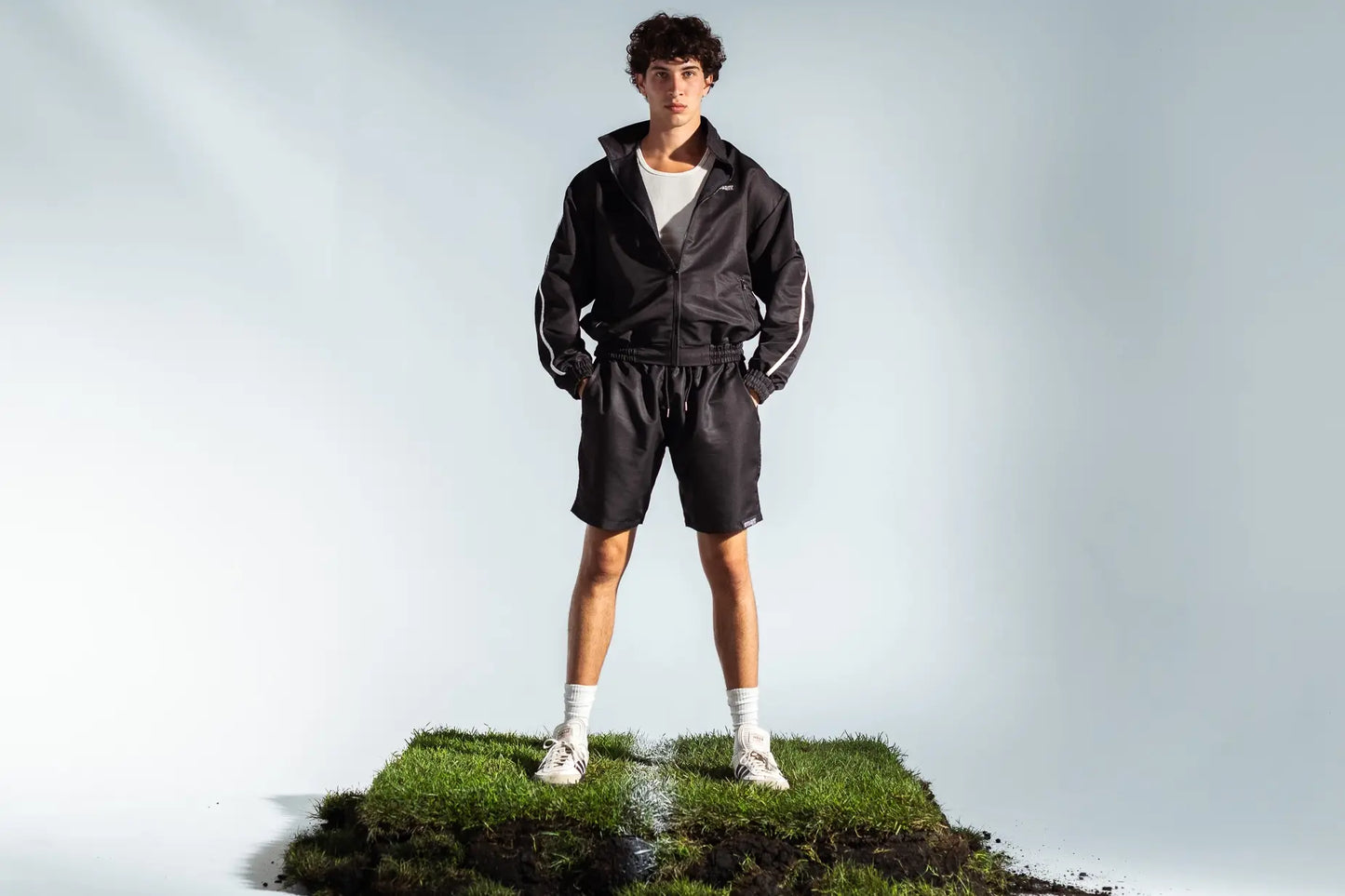 Model in full STMNT Wear black shorts and track jacket outfit standing on soccer turf with white sneakers
