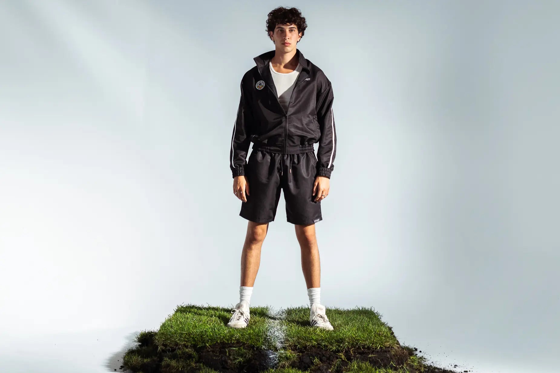 STMNT Wear black track jacket and shorts full outfit on model standing on soccer pitch turf cut-out inside a studio