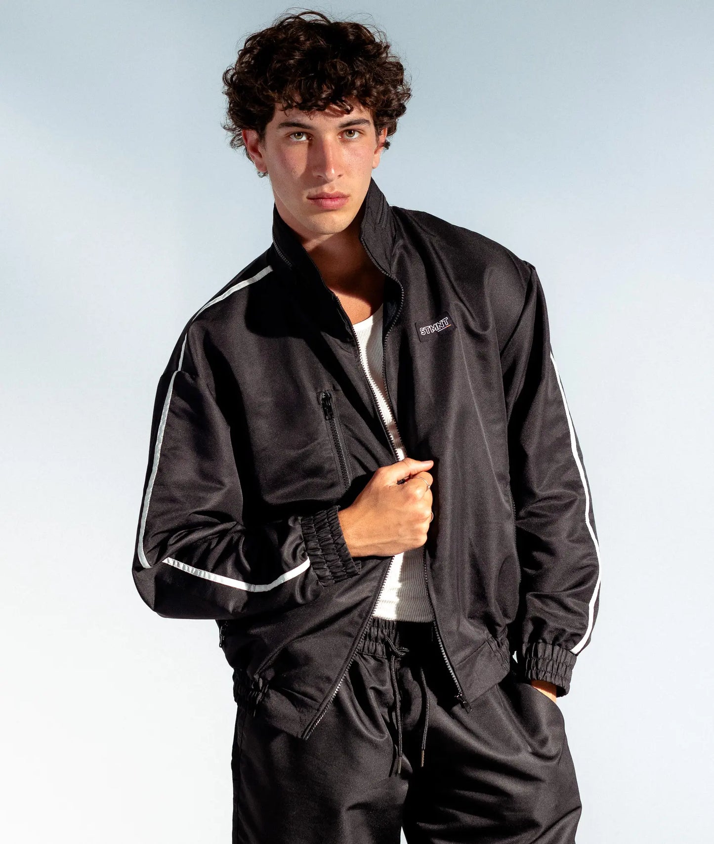 STMNT Wear black athletic track jacket with white sleeve stripes, front view on model over white tee