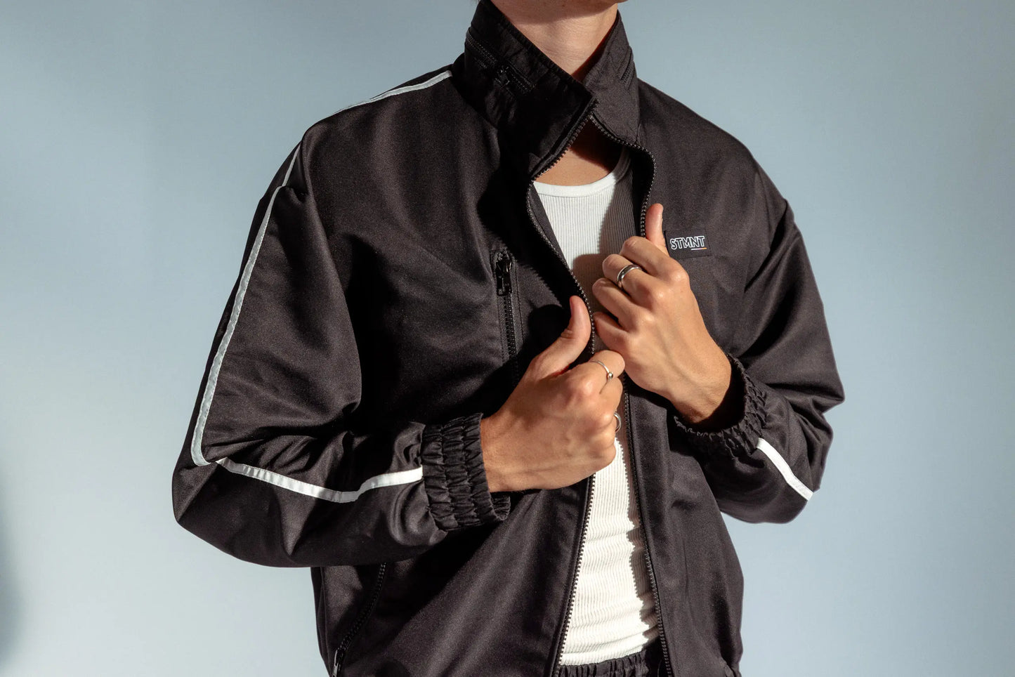 Close-up of STMNT Wear black track jacket showing chest logo, zip closure, and white piping detail