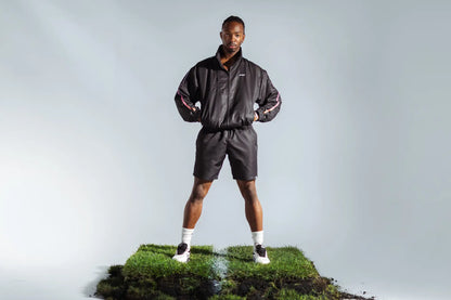 STMNT Wear First Edition black track jacket with orange-blue gradient accent stripes, full outfit on model having a wide stance on soccer turf cut-out in a studio