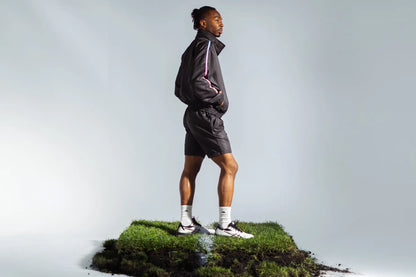 Model wearing STMNT Wear First Edition black athletic shorts and jacket standing on soccer pitch turf