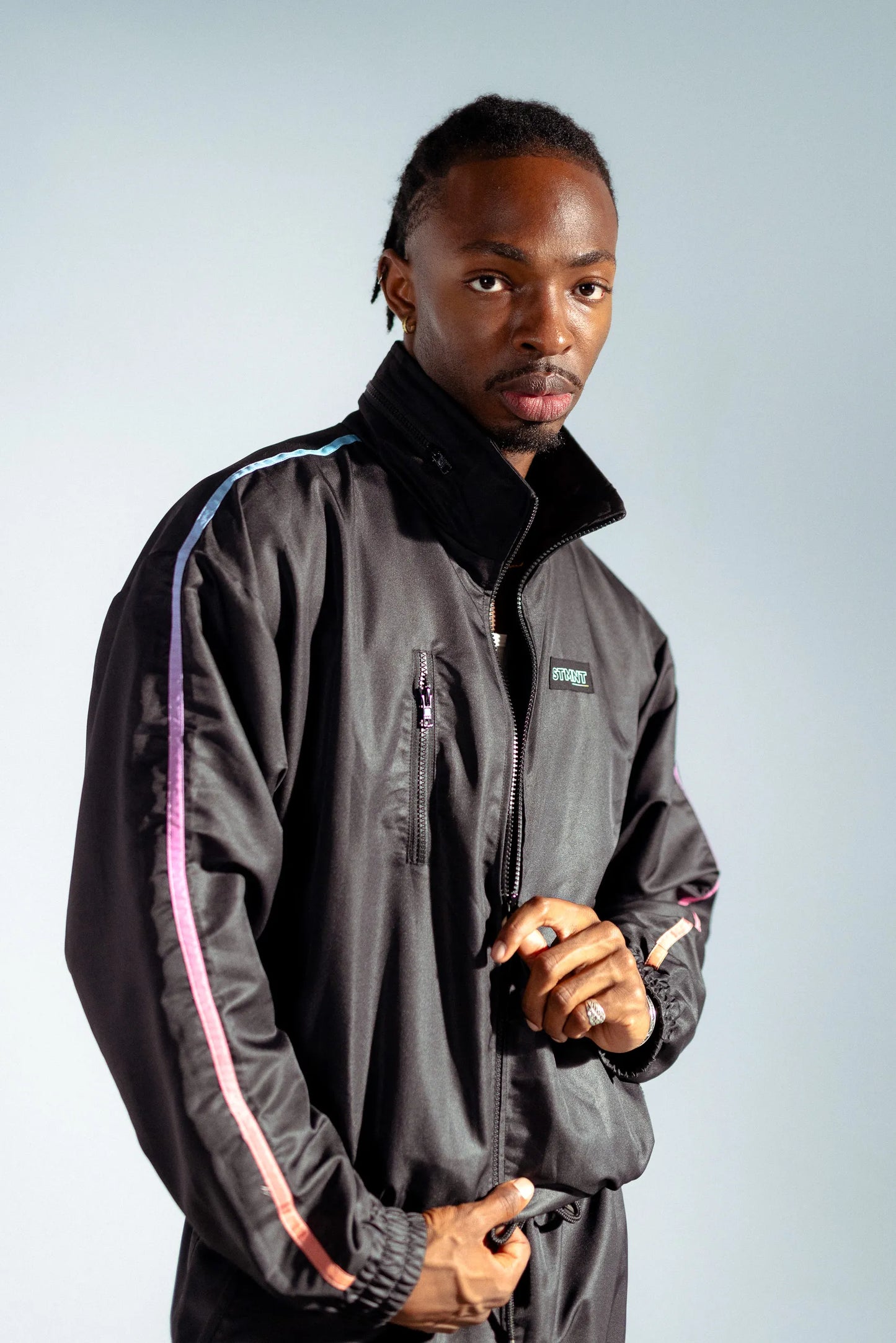 STMNT Wear First Edition black track jacket front view showing gradient rainbow sleeve stripe and STMNT logo patch