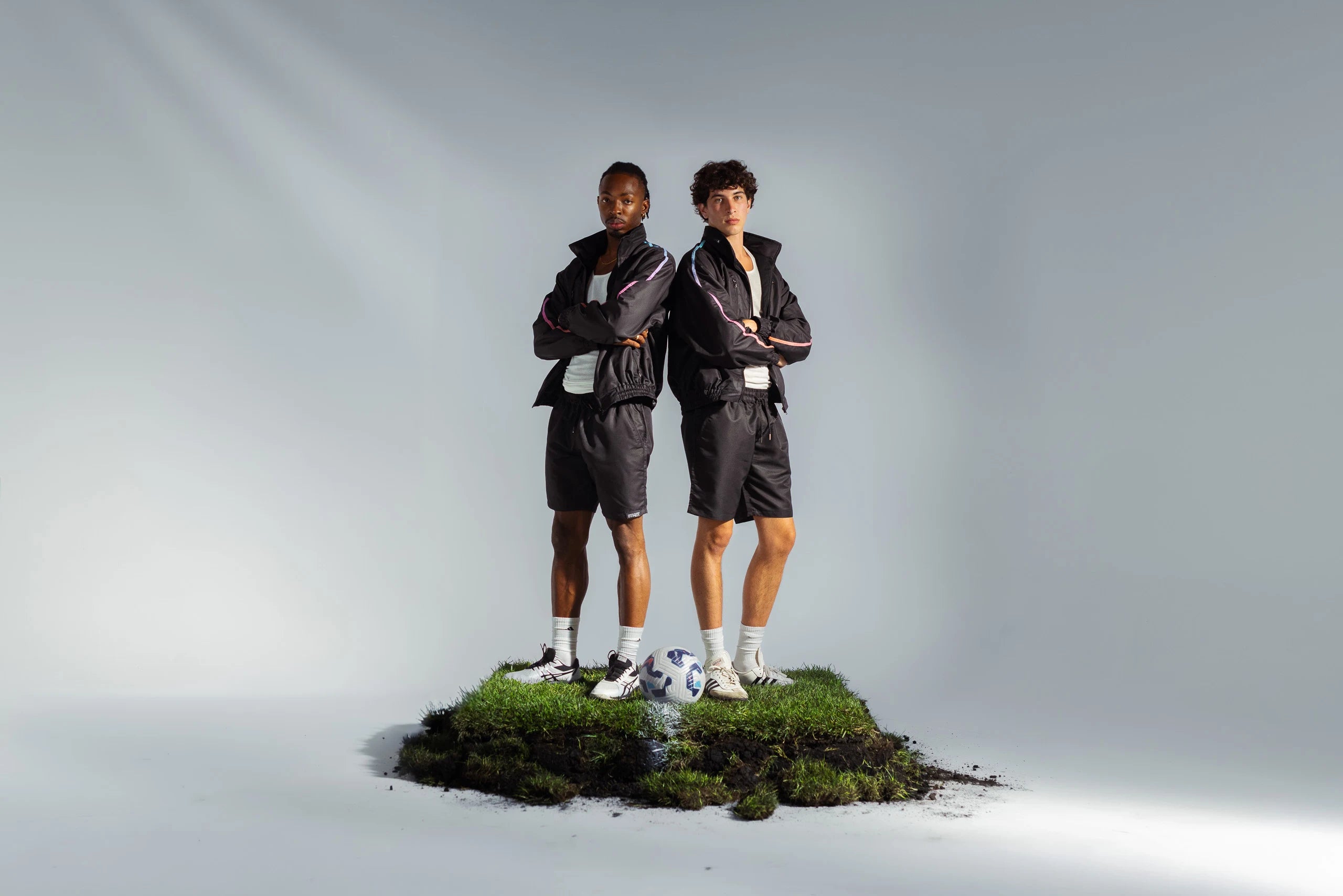 Two models standing on grass mound with soccer ball in matching STMNT Wear First Edition black track sets - modern athleisure set
