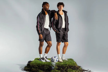 Two models wearing STMNT Wear First Edition Black Full Kit with soccer ball on grass turf cut-out in a studio, track jackets over white tees