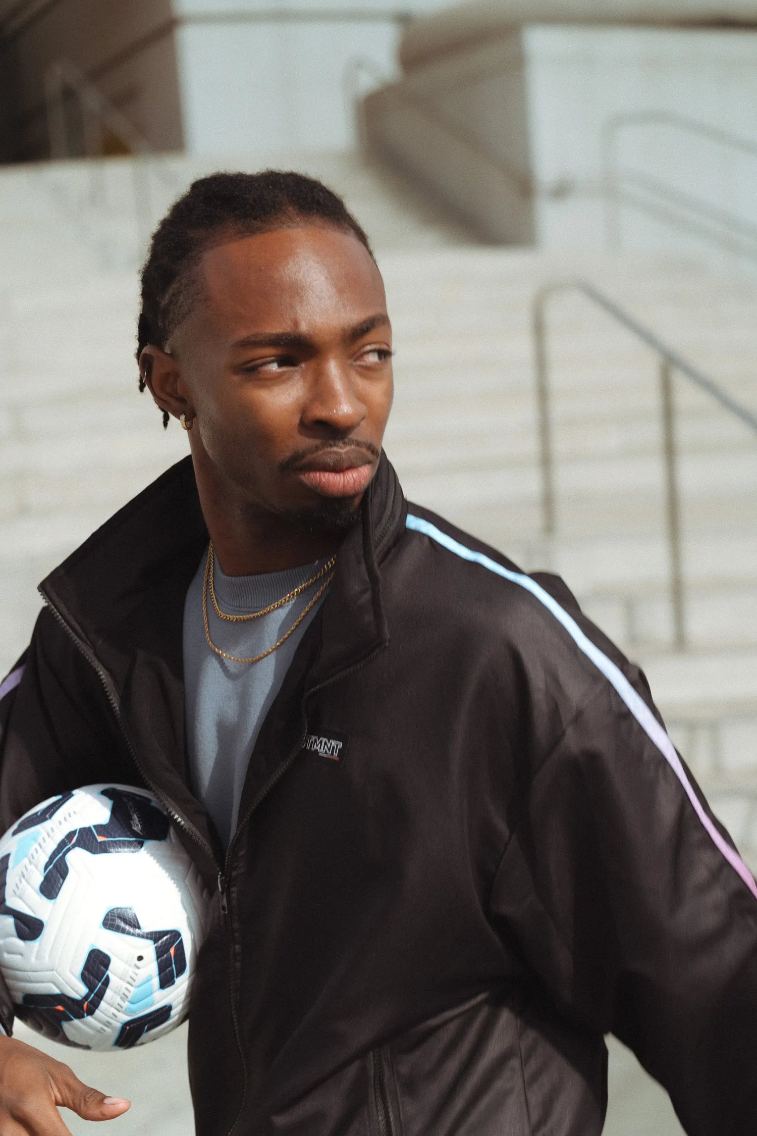 Model in STMNT Wear First Edition black track jacket with stripe detail holding soccer ball on concrete steps - soccer streetwear