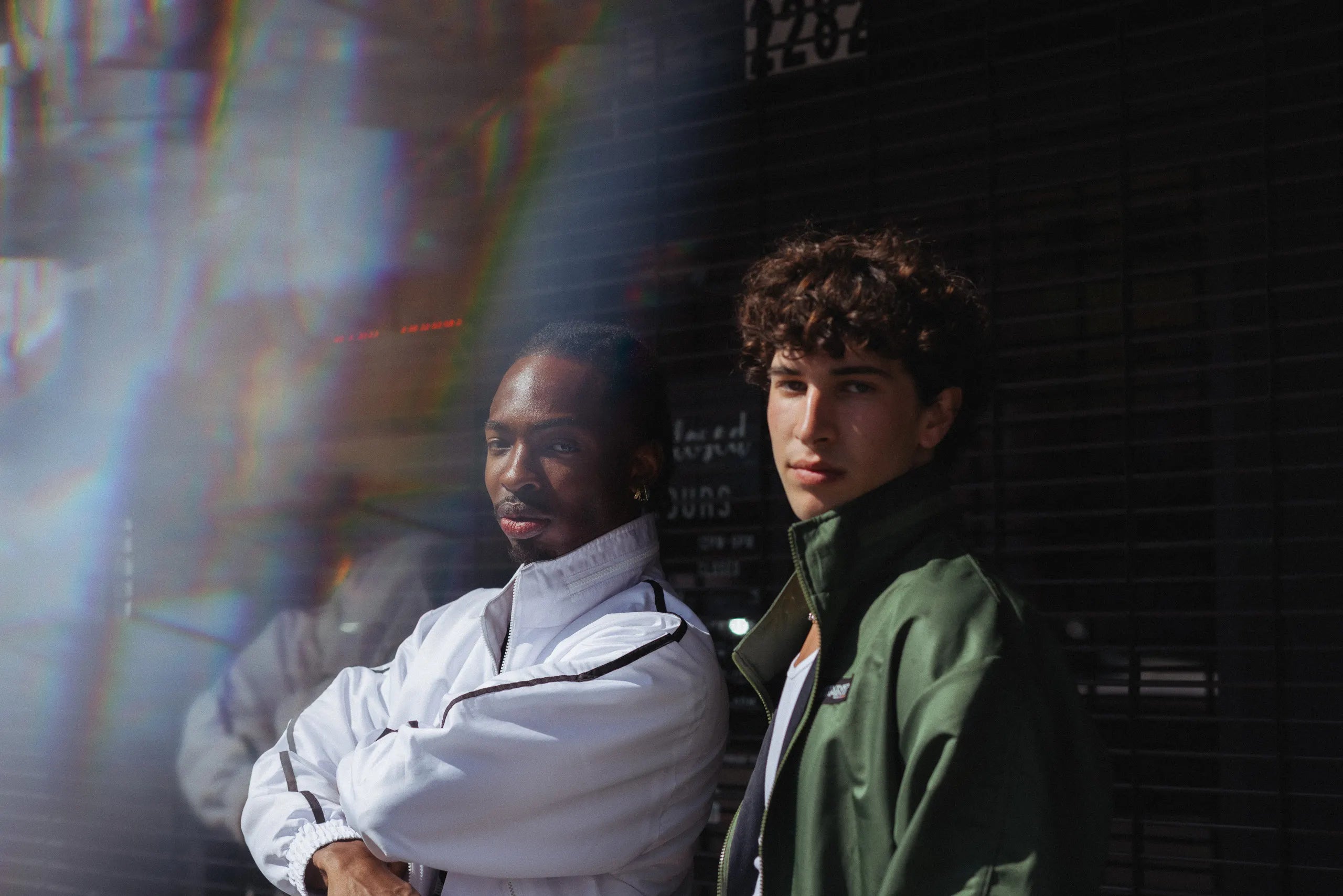 Two models in STMNT Wear white and green track jackets posing against dark urban storefront with lens flare