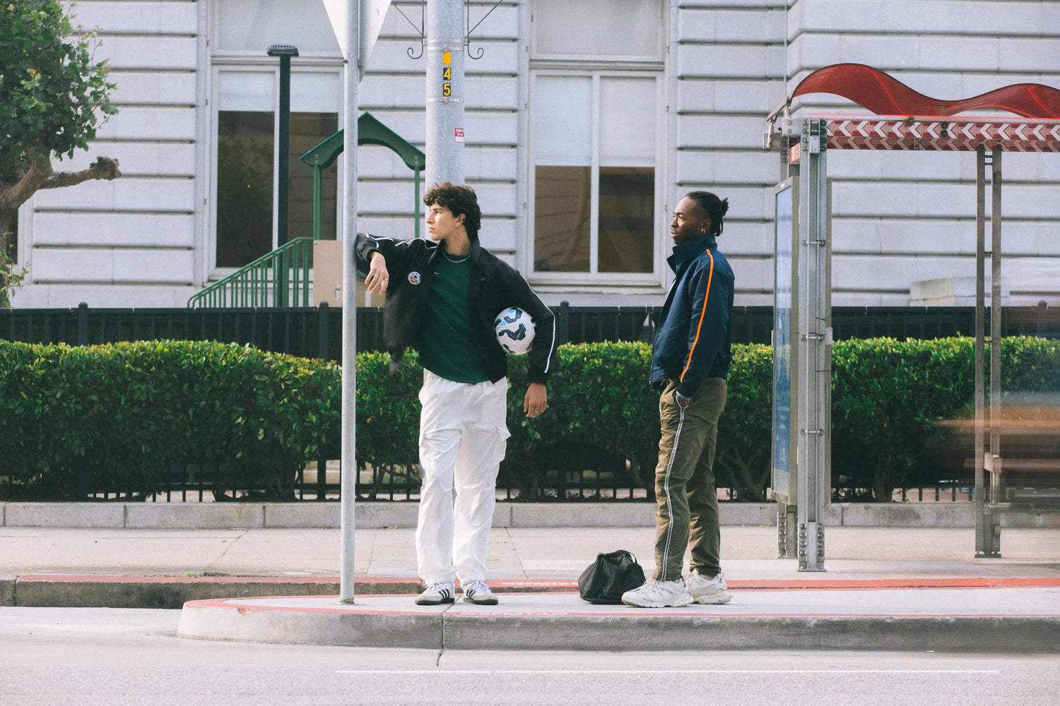 Two models in STMNT Wear track jackets with soccer ball at urban bus stop - athletic streetwear lookbook