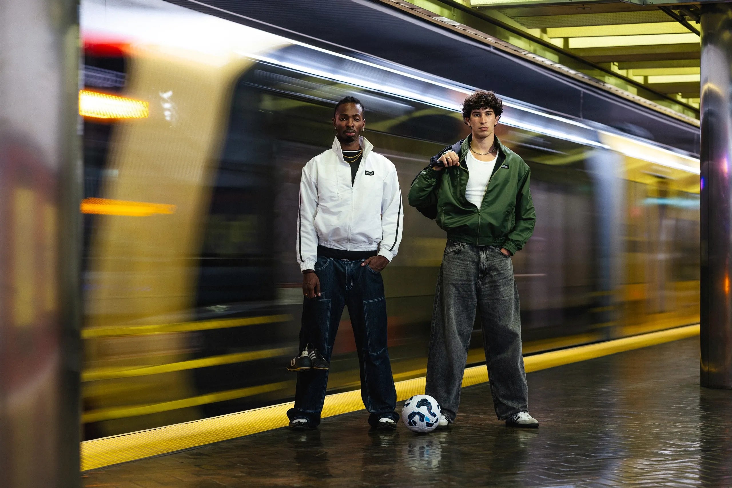 Two models in wearing jeans and STMNT Wear white and green jackets with soccer ball at subway platform - street meets game editorial